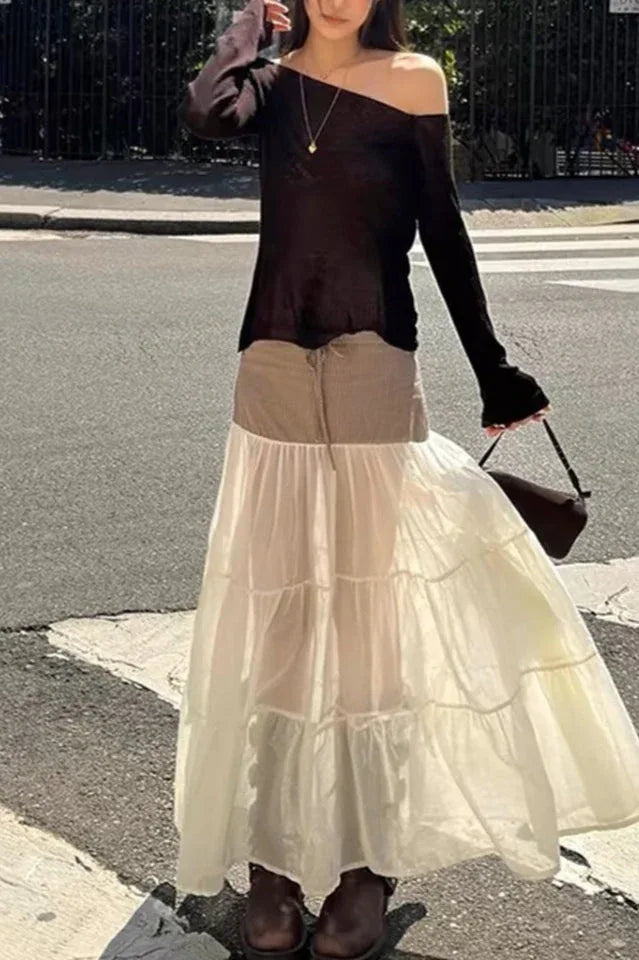 Model wearing a black off-the-shoulder top and a tiered maxi skirt with a brown pinstripe waistband, standing on a city street.
