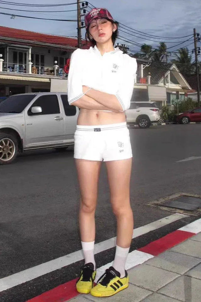 Woman standing outdoors wearing a white cropped polo shirt, white shorts, yellow sneakers, and a red and white checkered cap.