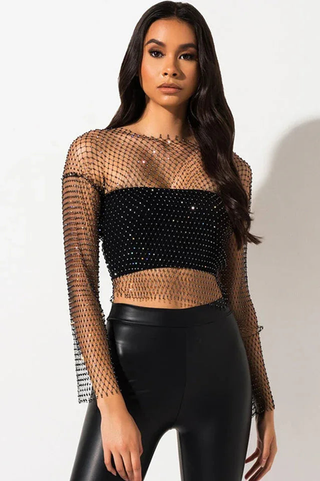 Woman wearing a black sheer long-sleeve crop top with sparkling embellishments over a black bandeau top, paired with black faux leather leggings.
