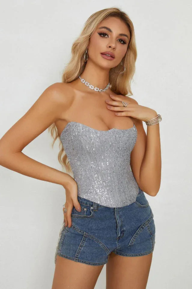 Woman wearing a silver sequined strapless corset top with a scalloped neckline and blue denim shorts, posing with hands on hips.