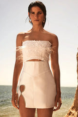 Woman wearing a white strapless feather and sequin crop top and a white high-waisted satin skirt with a cut-out detail, standing by the ocean.