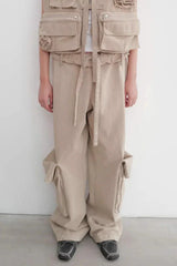 Person wearing a tan utility vest over a white tank top, paired with tan baggy cargo pants with prominent side pockets.