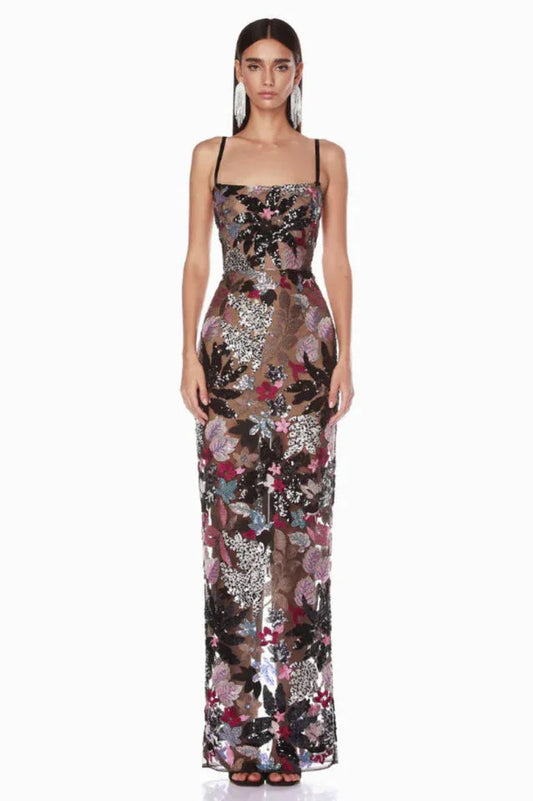 Woman wearing a floor-length black sheer gown with colorful floral appliqués and sequins, featuring spaghetti straps and a straight neckline.