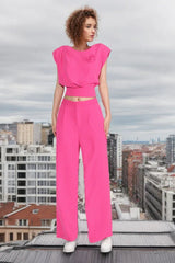 Woman wearing a bright pink two-piece set with a cropped top and wide-leg pants, standing on a rooftop with a city skyline in the background.