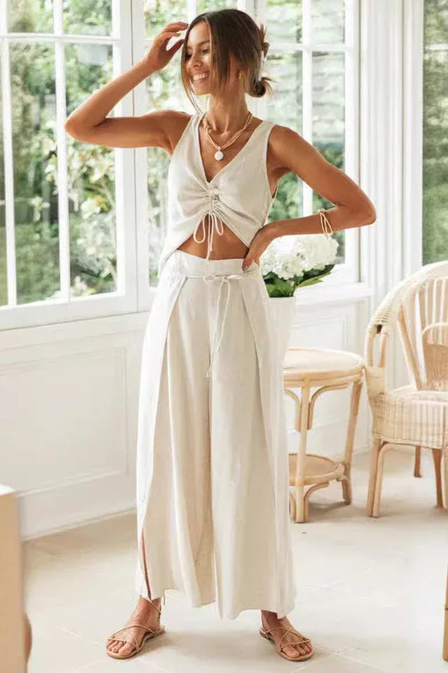 Woman wearing a cream linen-blend sleeveless v-neck ruched drawstring crop top and matching wide leg pants with side slits and tie waist.