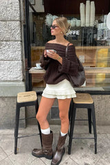 Model wearing a white pleated mini skirt with a brown off-the-shoulder sweater and brown boots.