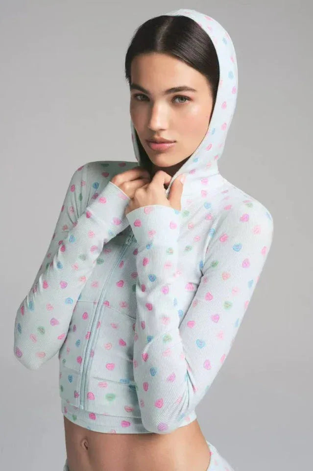 Close-up of a woman wearing a light blue cropped hoodie with heart prints, hands near her neck.