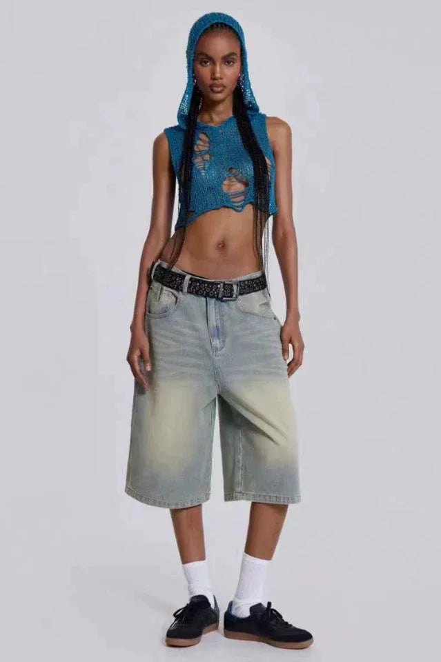 Woman wearing a blue distressed knit crop top with a hood and light wash wide-leg denim capri pants, a black belt, white socks, and black sneakers.