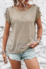 Woman wearing a tan short-sleeved t-shirt with button details on the shoulders and denim shorts.