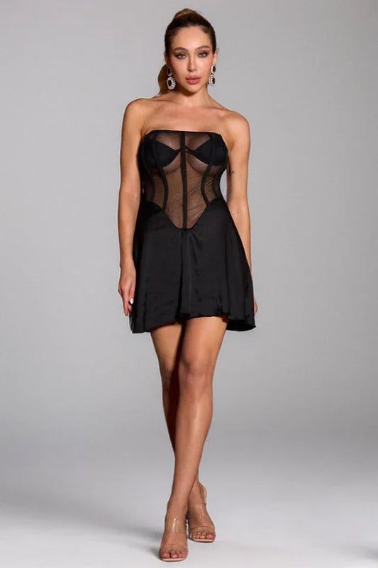 Woman wearing a black strapless mini dress with a sheer corset bodice and a full skirt, standing against a neutral background.