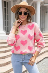 Woman wearing a pink crew neck sweater with a textured pink heart pattern, light blue jeans, a hat, and sunglasses.