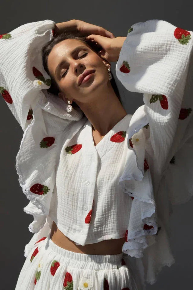Woman posing with her arms raised, showcasing the ruffled sleeves and strawberry print of the white blouse.