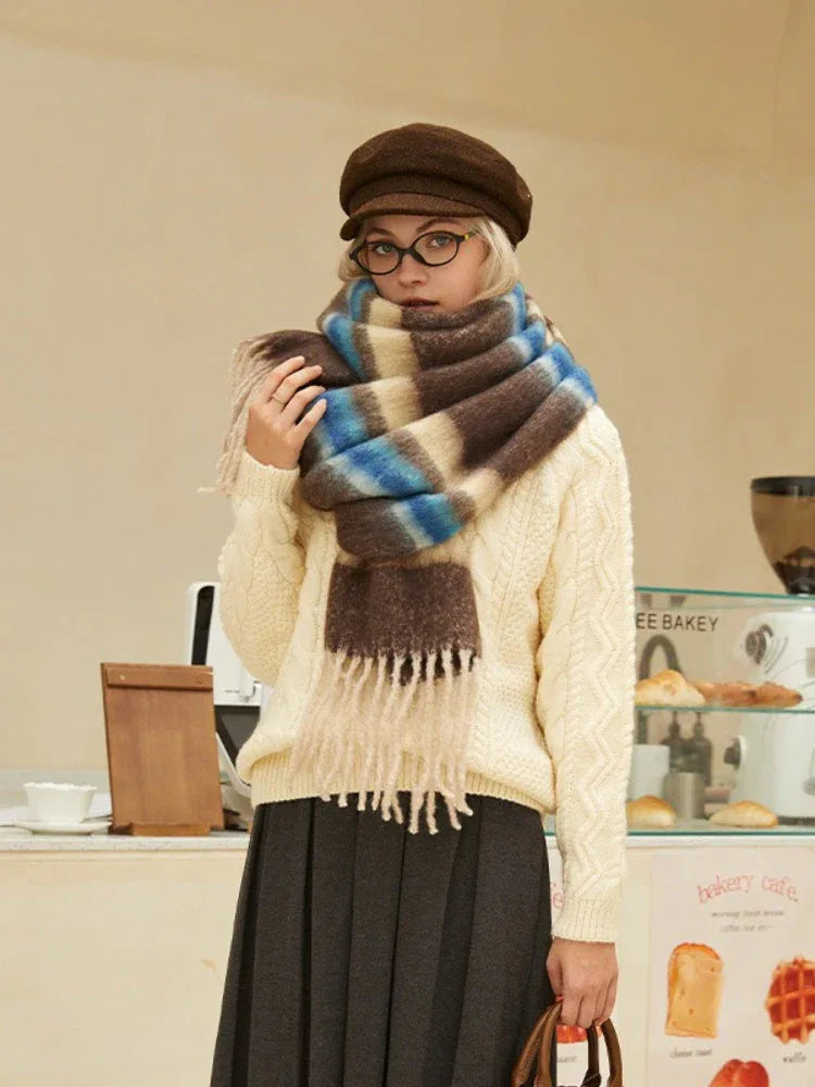 Model wearing a cream cable knit sweater and a brown plaid scarf with cream, blue, and brown stripes. She is also wearing a brown flat cap and round glasses, and a dark grey pleated skirt.