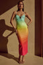 Full-length image of a model wearing a teal, yellow, orange, and pink ombre sequined midi dress with spaghetti straps and a dragonfly embellishment at the bust, standing against a tan wall with shadows.