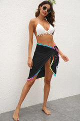 Woman wearing a white bikini top and a black sarong with colorful pom-pom trim tied at the hip.