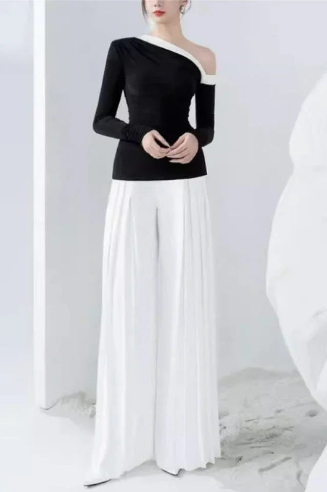 Full-body shot of a model in a black asymmetrical top and white wide-leg pleated pants, hands clasped.