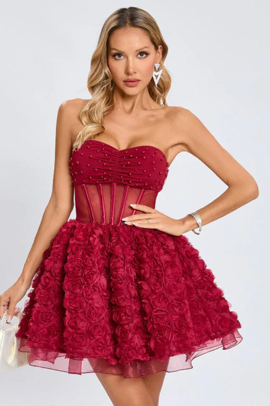 Woman wearing a short, strapless, deep red dress with a ruched and beaded bodice, a sheer corset waist, and a full skirt decorated with 3D rose appliques.