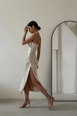 Woman in a champagne satin slip dress with a cowl neckline and high slit, posing with her hand on her forehead.