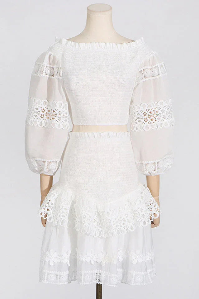 Mannequin displaying a white off-the-shoulder smocked crop top with sheer lace sleeves and a white tiered lace skirt