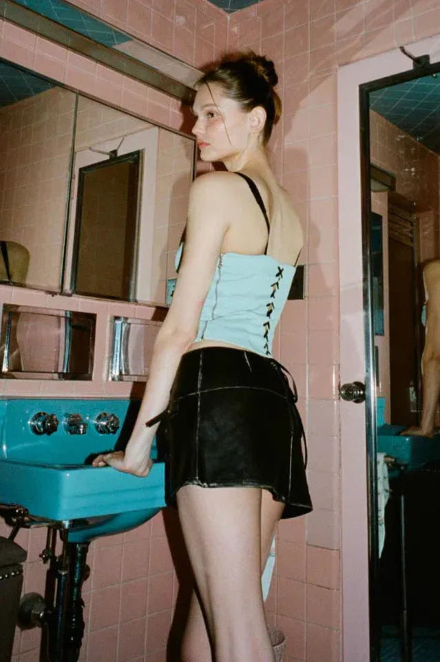 A young woman stands in a pink tiled bathroom, showing the back of a light blue cropped top with black trim and a black mini skirt.