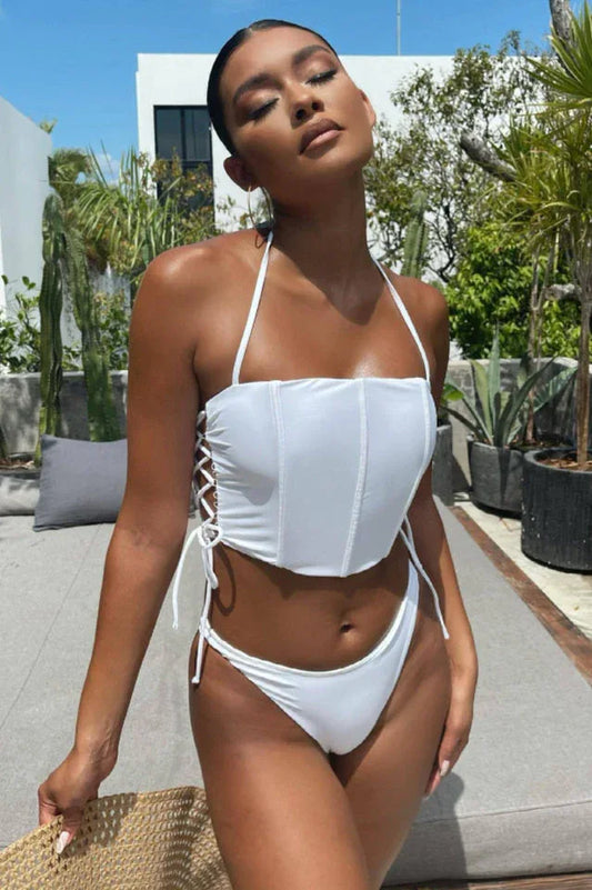 Woman in a white two-piece swimsuit with a corset-style top and lace-up sides, posing outdoors with lush greenery in the background.
