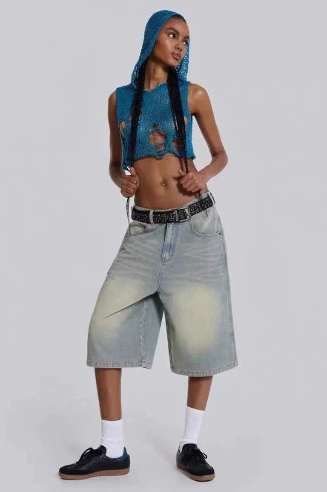 Woman adjusting the sleeves of a blue distressed knit crop top with a hood, paired with light wash wide-leg denim capri pants, a black belt, white socks, and black sneakers.