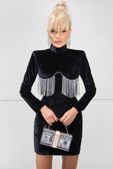 Woman wearing a black velvet mini dress with a high neckline, long sleeves, and sparkling fringe detail across the bust, holding a money-themed clutch.