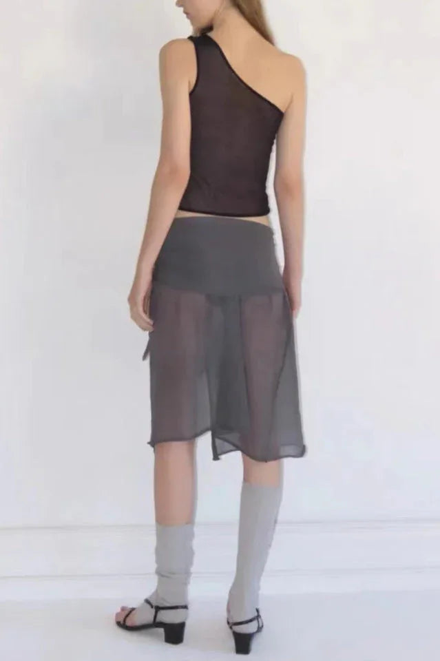 Full body back view of a woman wearing a black asymmetrical sheer knit top and a sheer grey skirt.