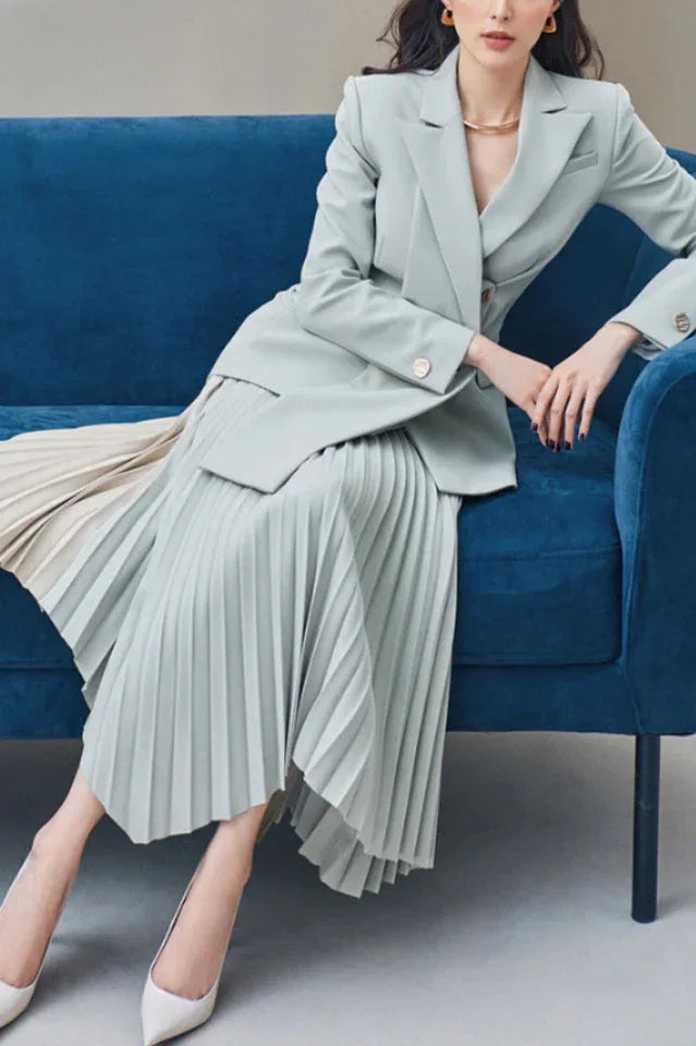 Woman wearing a light blue blazer and asymmetrical pleated skirt, sitting on a blue couch.