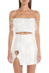 Front view of a woman wearing a white strapless feather and sequin crop top and a white high-waisted satin skirt with a cut-out detail.