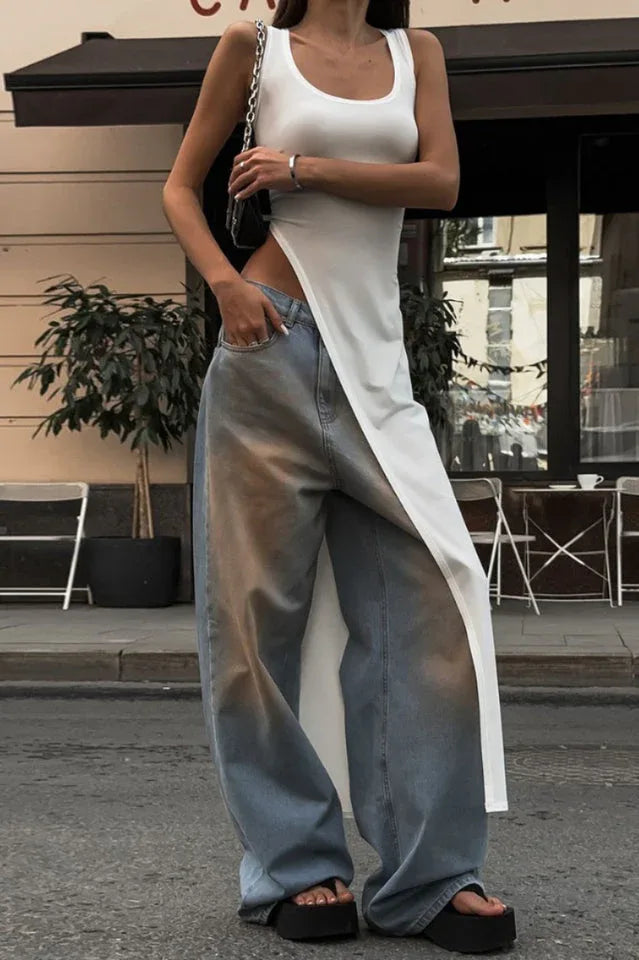 Close-up of a woman wearing a white sleeveless maxi dress with a high slit over light blue wide-leg jeans, showing the outfit details
