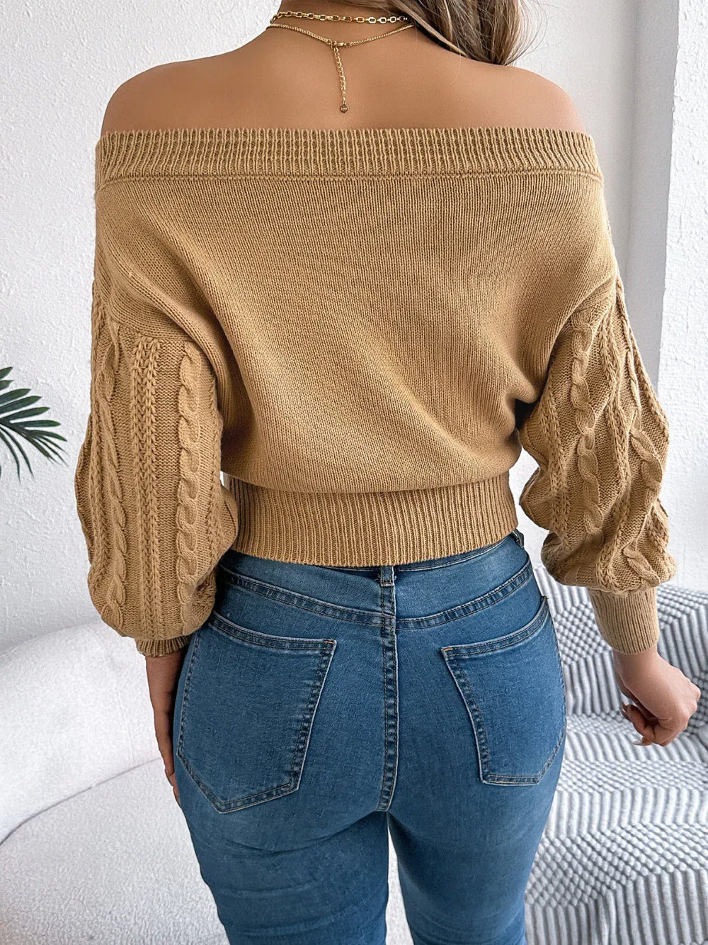 Close-up of a woman's torso wearing a tan off-the-shoulder cable knit cropped sweater, blue jeans, and jewelry.