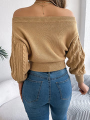Close-up of a woman's torso wearing a tan off-the-shoulder cable knit cropped sweater, blue jeans, and jewelry.