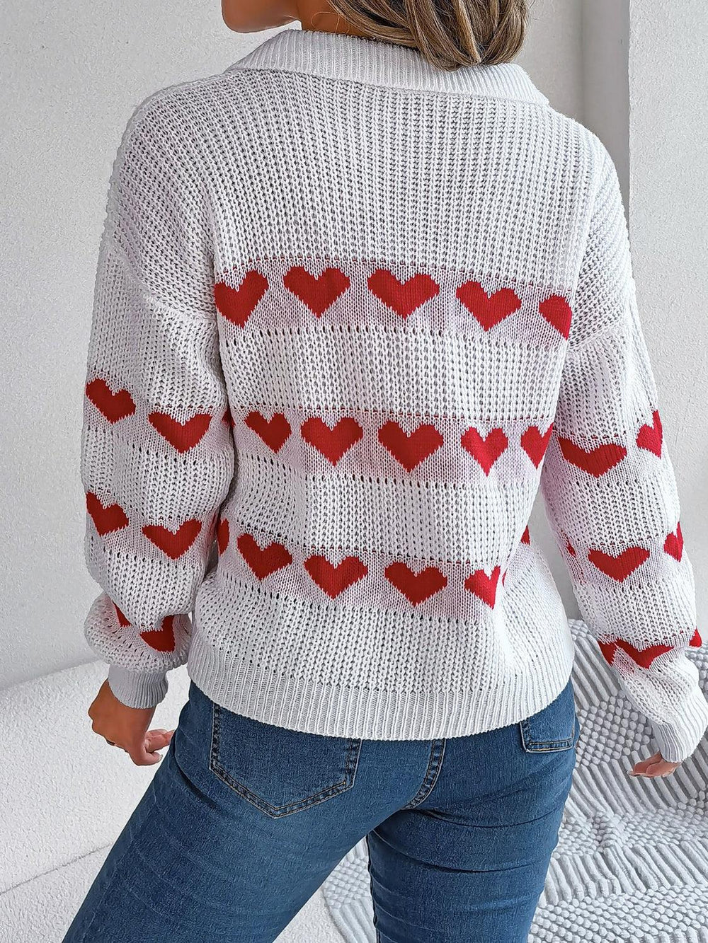 Back view of a woman wearing a white knit sweater with red heart pattern and blue jeans