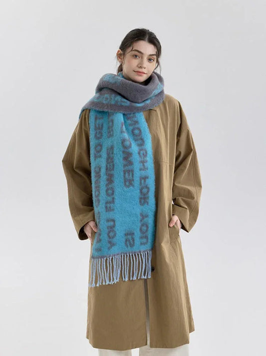 A woman models a blue and gray knit scarf with text print and fringe detail, worn over a tan trench coat.