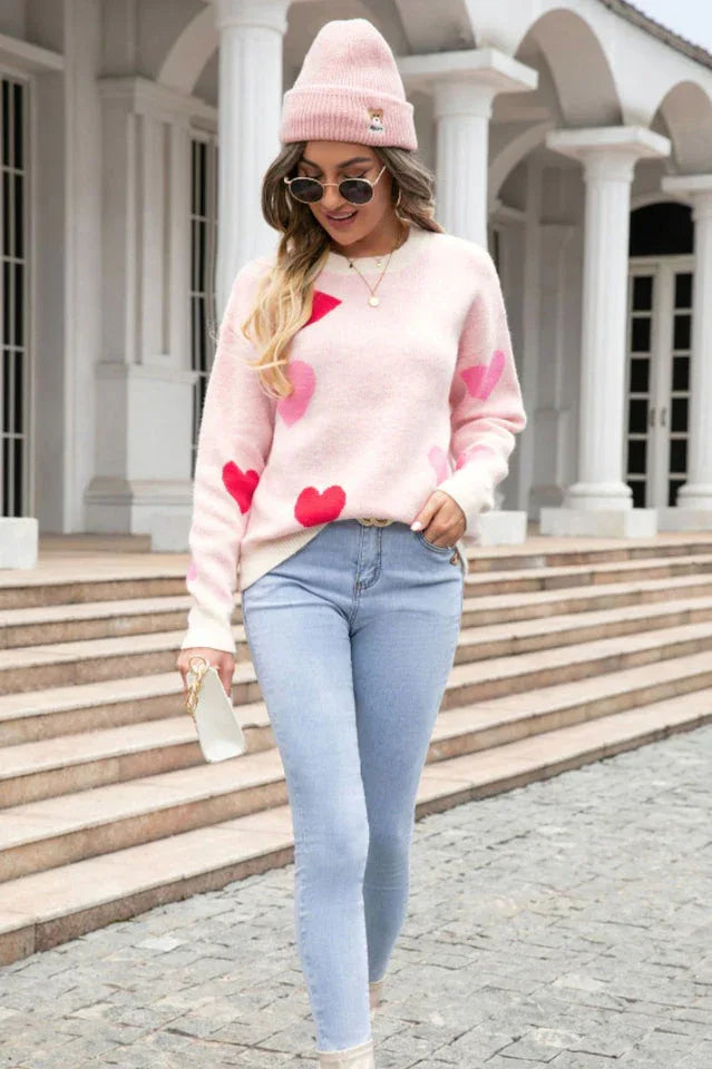 A woman wearing a pink heart print sweater, pink beanie, and light wash jeans, walking.