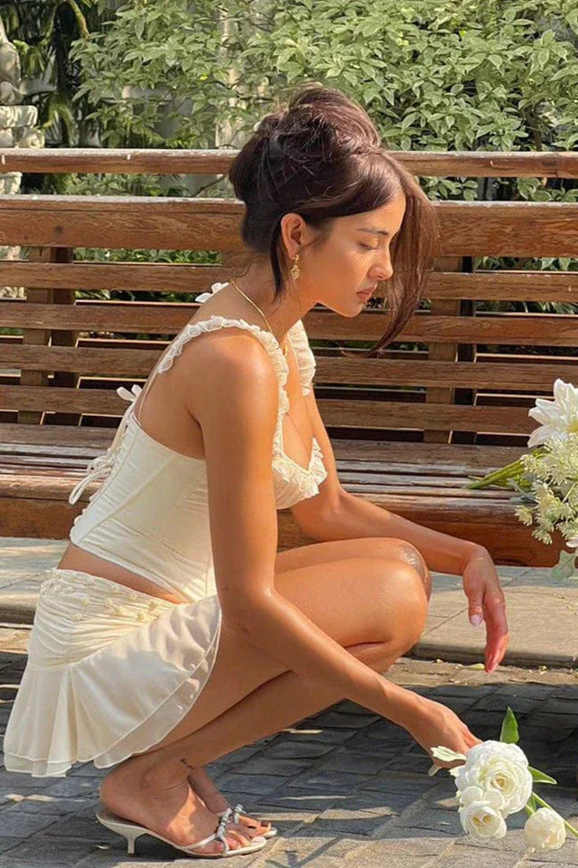 Woman in a white corset top and skirt crouching down to pick up flowers.
