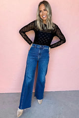 Full-length shot of a woman wearing a black sheer long-sleeve top with an all-over bow print, layered necklaces, and wide-leg blue jeans, standing against a pink background.