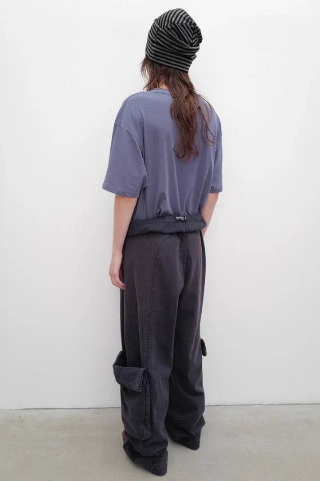 Rear view of a person wearing a purple graphic t-shirt, dark wash baggy cargo pants with prominent side pockets, and a striped beanie.