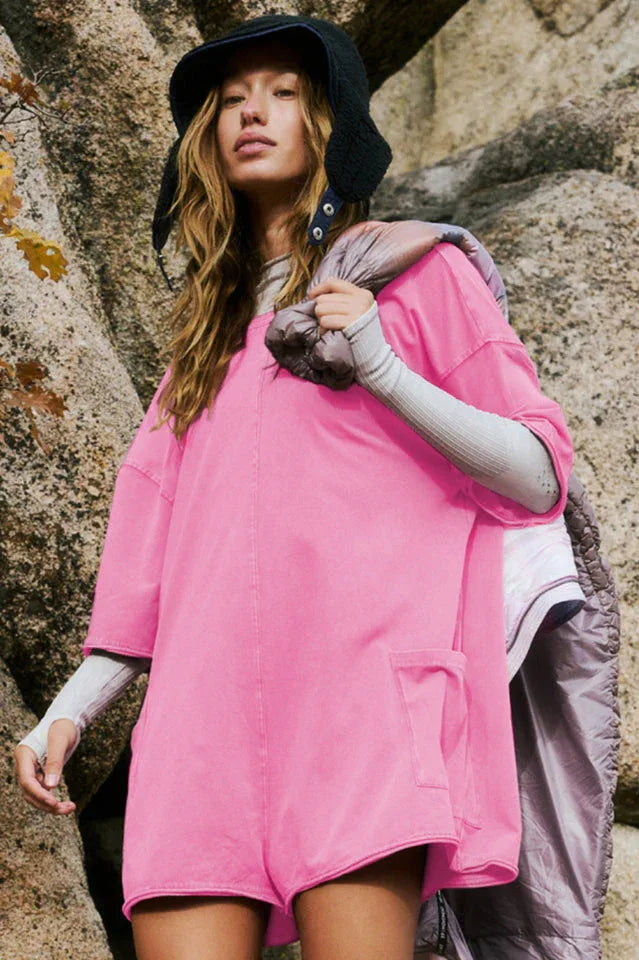 Woman wearing a bright pink oversized tunic with short sleeves and pockets, layered with a grey long-sleeve top and a dark hat, outdoors.