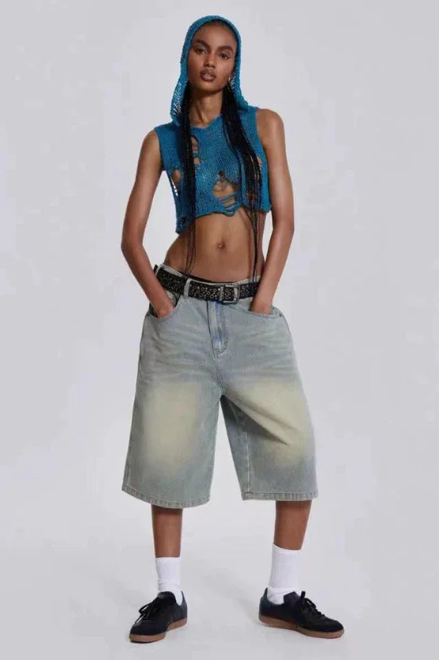 Woman with hands in pockets wearing a blue distressed knit crop top with a hood and light wash wide-leg denim capri pants, a black belt, white socks, and black sneakers.