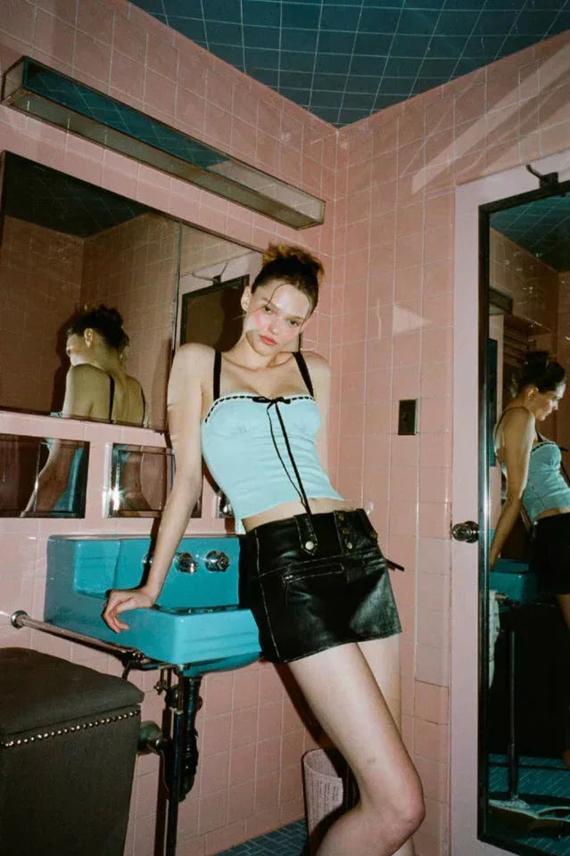 A young woman poses in a pink tiled bathroom, wearing a light blue cropped top with black trim and a black mini skirt.