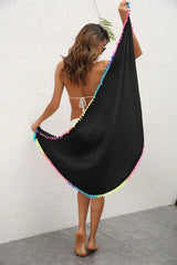 Rear view of a woman holding a black sarong with colorful pom-pom trim outstretched.
