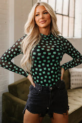Woman wearing a black long-sleeved mesh bodysuit with a green shamrock print and black denim shorts, posing with hands on hips.