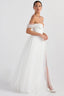 Model in a white off-the-shoulder wedding dress with a high slit, standing against a white background.