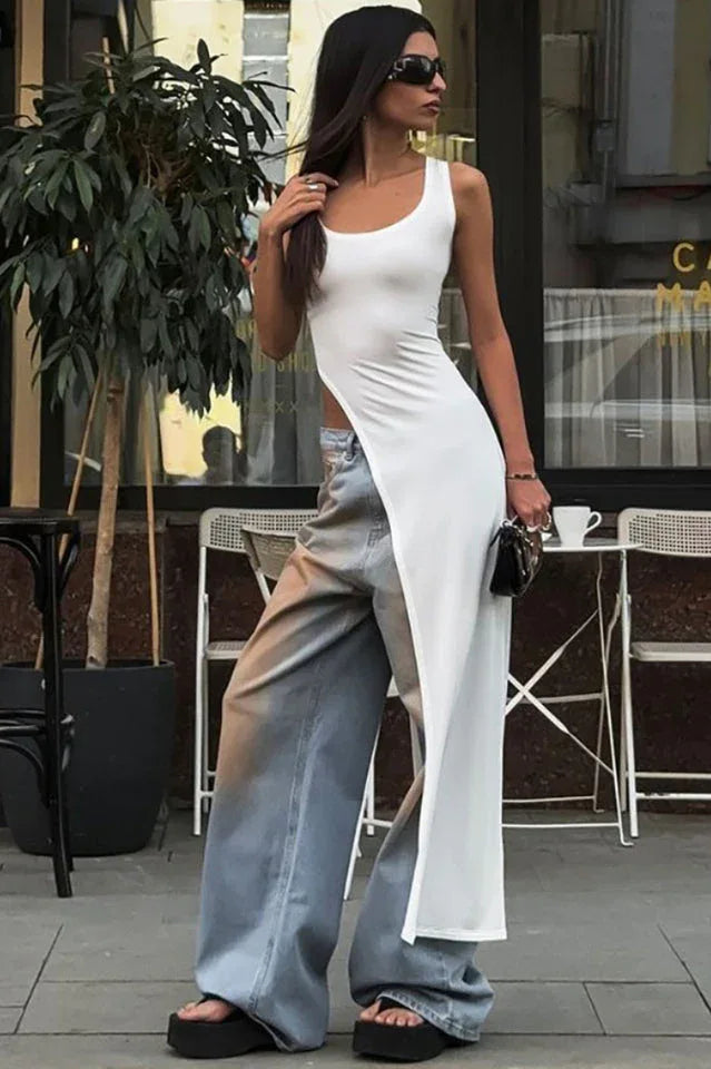 Woman wearing a white sleeveless maxi dress with a high slit over light blue wide-leg jeans and black platform sandals outdoors