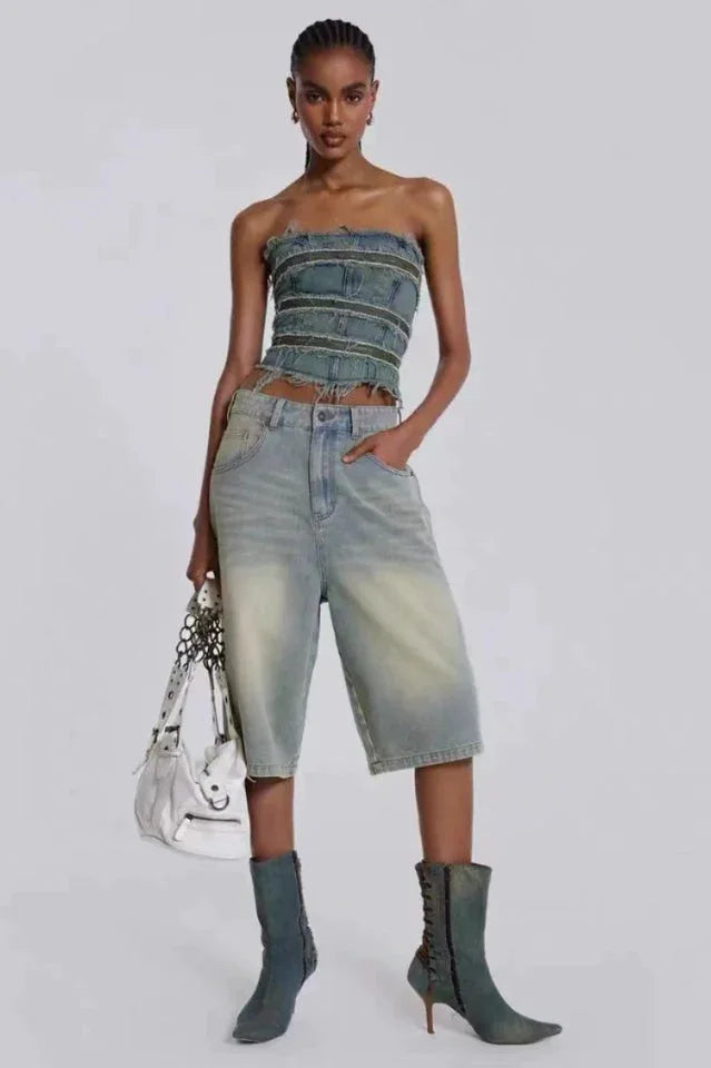 Woman wearing a denim strapless corset top with frayed edges and light wash wide-leg denim capri pants, paired with teal heeled boots and a white handbag.