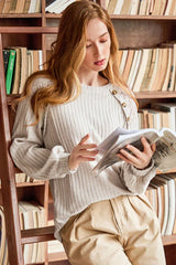 Woman wearing a cream ribbed knit sweater with lace shoulder detail and tan pants, reading a book in front of a bookshelf.