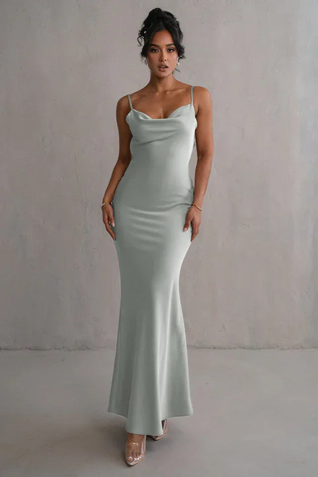 woman wearing a light green satin floor-length mermaid dress with spaghetti straps and a cowl neckline, standing against a grey wall