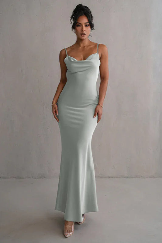 woman wearing a light green satin floor-length mermaid dress with spaghetti straps and a cowl neckline, standing against a grey wall